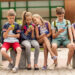 primary education, friendship, childhood, technology and people concept - group of happy elementary school students with smartphones and backpacks sitting on bench outdoors