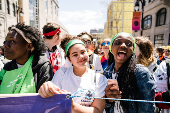 Smiling young people at the front of a group who is marching in the streets.