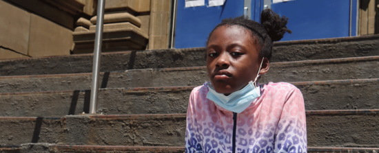 Une fille noire avec un masque autour du cou est assise sur les marches devant une école.